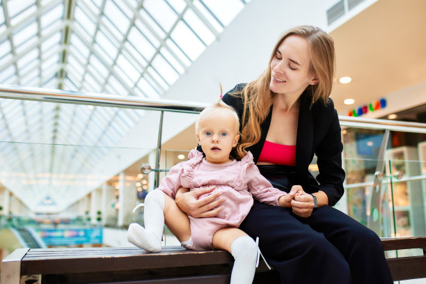 一位年轻漂亮的母亲带着一个小婴儿坐在购物中心的长椅上。穿着粉色衣服的妈妈和女儿在购物时拥抱、玩耍、放松和享受乐趣。家庭周末在购物中心。图片下载