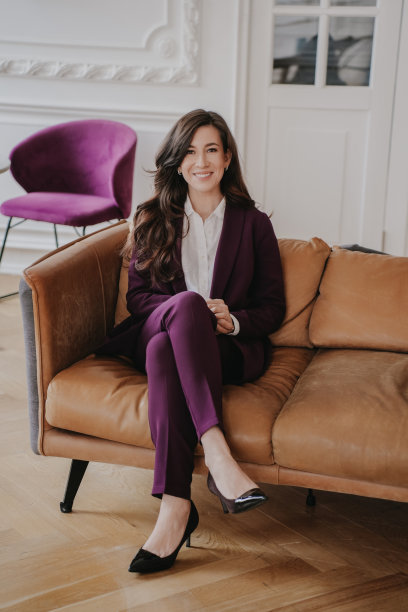 办公室里穿着紫色套装坐在皮沙发上的开朗的女经理的垂直照片。深色皮肤的美国女商人，因获得成功而咧嘴笑。商业和金融。图片下载