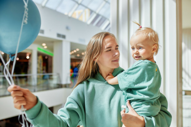 母亲的特写肖像，怀里抱着婴儿和彩色气球。年轻漂亮的妈妈带着一个穿着同样绿色衣服的小女儿在商场里散步，玩得很开心。幸福的家庭。图片下载