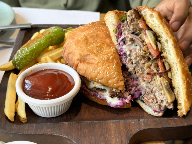 特写照片中，一个认不出来的人拿起半个汉堡、西红柿、泡菜、生菜放在芝麻面包里，放在木板上，配上酥脆的薯条，西红柿小杯，抬高视野，聚焦在前景上图片下载