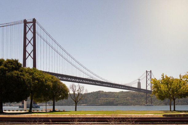 4月25日葡萄牙里斯本大桥的日间、地面水平景观照片，大桥横跨一条河流，前景是绿树，背景是群山，天空湛蓝图片下载