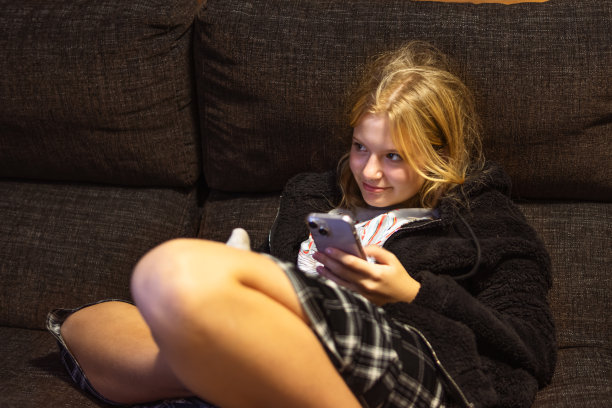 一个外表浅色的十几岁女孩坐在深色的沙发上，手里拿着一部手机。高质量照片图片下载