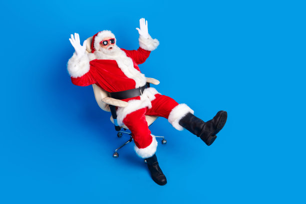 老人的全尺寸照片惊讶地举起手扶手椅穿着时尚的圣诞老人服装外套孤立的蓝色背景图片下载