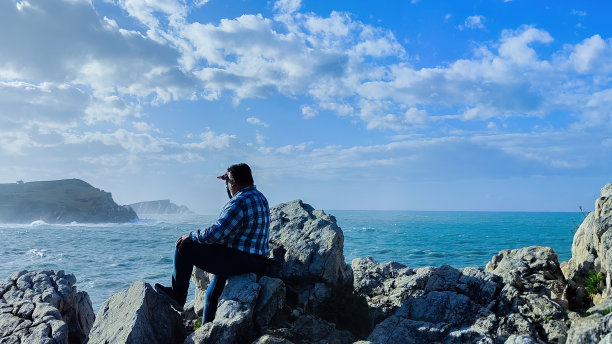 男人坐在悬崖上的岩石上，海浪挥手，用他的手在海上的宁静与水平的照片复制空间图片下载