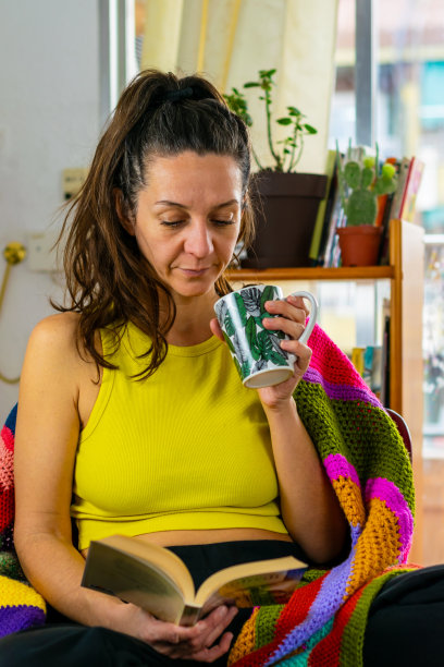 一个女人用温暖的毯子享受阅读，同时从一个杯子里喝，气氛是温暖的，有色彩，垂直的照片复制空间图片下载