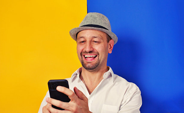 照片肖像中快乐的男人双手拿着手机，隔在黄色和蓝色，两种颜色的背景上，笑容灿烂，感到幸福图片下载