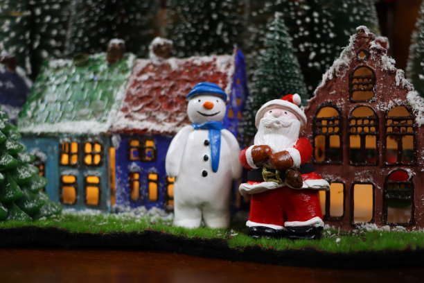 圣诞老人和雪人的特写图片在风景优美的圣诞村展示，人造草坪上的人造塑料云杉圣诞树和羊毛雪围绕着一排被照亮的自制粘土房屋，重点在前景图片下载