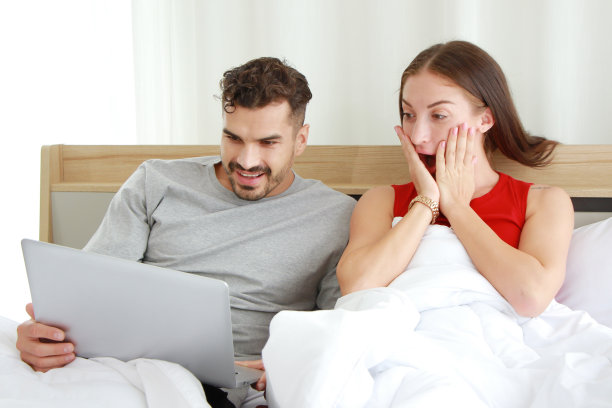 微笑的高加索夫妇是令人惊讶的，在卧室里用笔记本电脑在社交媒体上看在线电影很有趣。在家工作和度假的生活方式图片下载