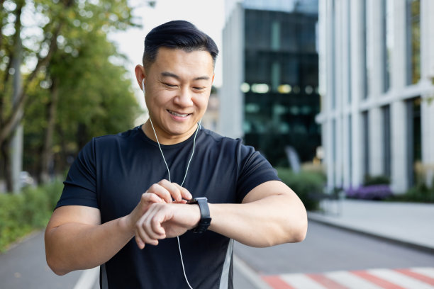 特写照片。年轻的亚洲男运动员戴着耳机站在城市街道上看手表图片下载