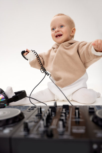 一个穿着米色连帽衫、面带微笑的可爱小孩坐在地板上，戴着dj耳机和dj混音板。音乐和乐趣。孤立在白色背景上。图片下载