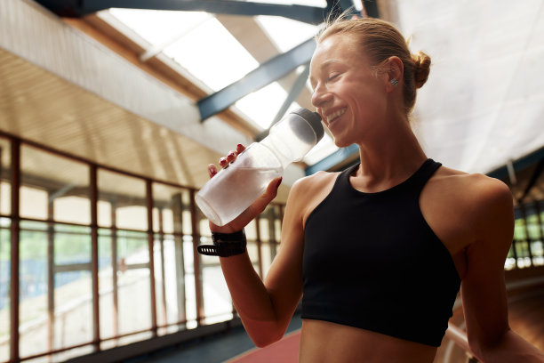年轻的运动员女孩在跑道上锻炼后正在休息。一个身穿黑色运动服、身材苗条、运动健将的金发女郎从运动瓶里喝水。运动和娱乐的概念。半身的照片。图片下载