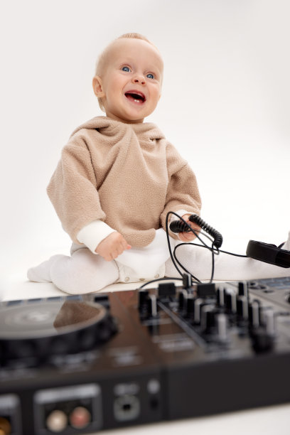 一个穿着米色连帽衫、面带微笑的可爱小孩坐在地板上，戴着dj耳机和dj混音板。音乐和乐趣。孤立在白色背景上。图片下载