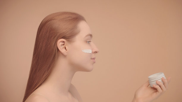 侧视图:一位脸颊上涂着面霜的年轻女子，手里拿着一瓶面霜。图片下载