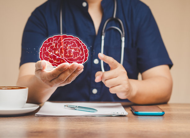 医生坐在医院的桌子旁，手里拿着一个红色的大脑符号。图片下载