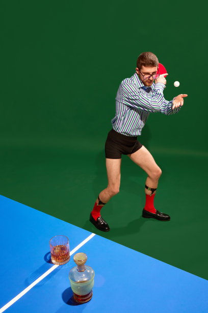 前视图。有趣的创意照片与男子穿着衬衫和内衣打乒乓球与威士忌玻璃杯在绿色背景。图片下载