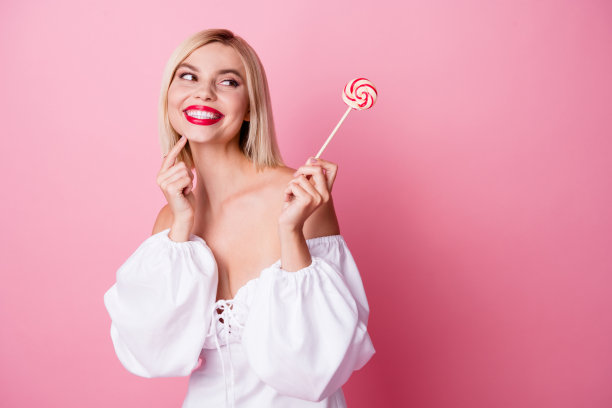 照片中开朗可爱诱人的女孩穿着时髦的白色衣服，吃着棒棒糖，在粉红色的背景上看起来空无一物图片下载