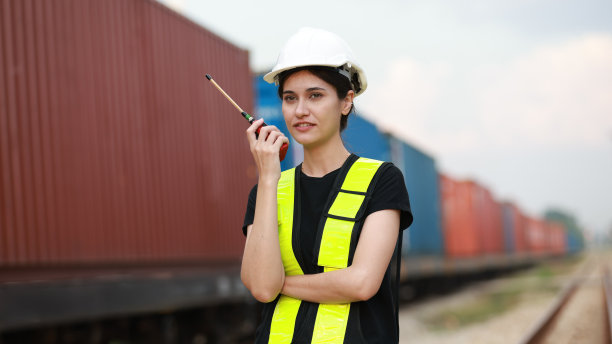 照片中的美女工程师身穿绿色安全背心，头戴白色安全帽，手持对讲机在火车站工作图片下载
