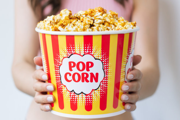 一位身穿粉色衣服的年轻女子面前拿着一大颗又甜又好吃的焦糖爆米花，装在一个鲜亮漂亮的包装里，上面有红黄相间的条纹。特写照片图片下载