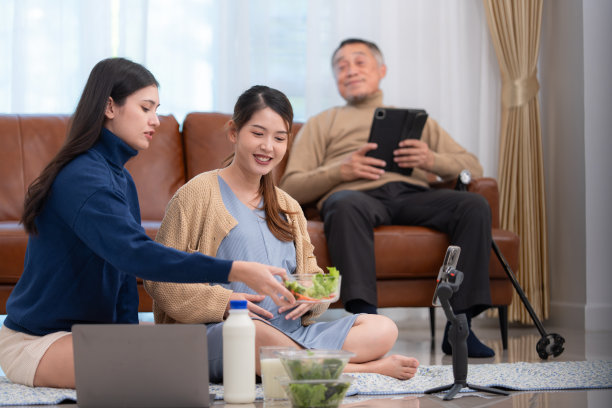 一个怀孕的亚洲妇女和她的妹妹坐在地板上。社交媒体直播销售孕妈妈产品和膳食，背景是一位父亲在沙发上放松。图片下载