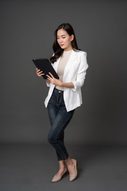 长发亚洲女高管，公司老板的全身照片。通过平板电脑做生意照片摄于工作室，背景为灰色，穿着白色西装、长牛仔裤和鹿皮鞋。图片下载