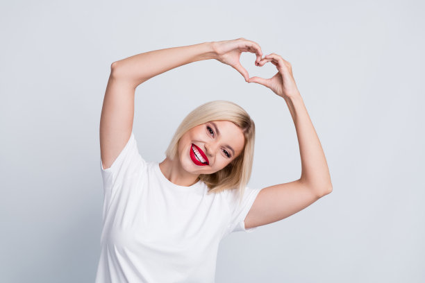 照片中开朗、漂亮、甜美、闪亮的女孩穿着时髦的衣服，手臂高举在头顶，在灰色背景上展示了孤立的心脏形象图片下载