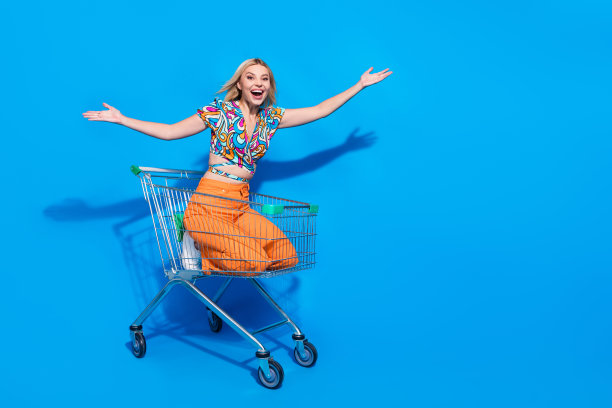 正面可爱的女孩坐在购物车手掌展示产品的全长照片，在蓝色背景上孤立的空白空间图片下载