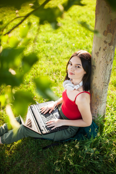 深棕色头发的女孩戴着白色耳机在公园的树下使用笔记本电脑图片下载
