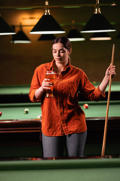 酒吧台球桌旁，年轻白人女子手持球杆，手持啤酒的照片。图片下载