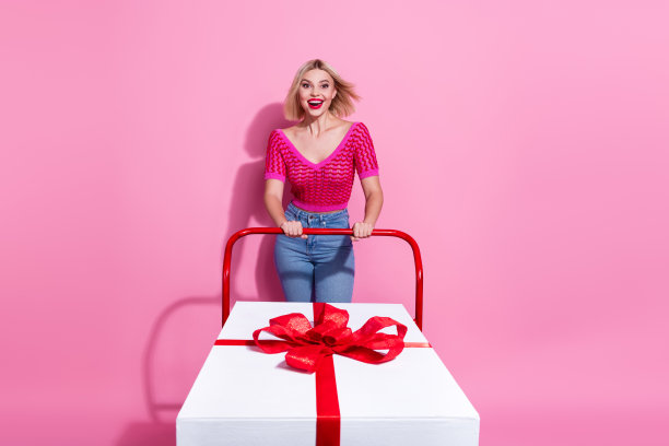 全身照片欣喜若狂的女人飘动的头发拿着购物车与大礼物惊讶的凝视孤立在粉红色的背景图片下载