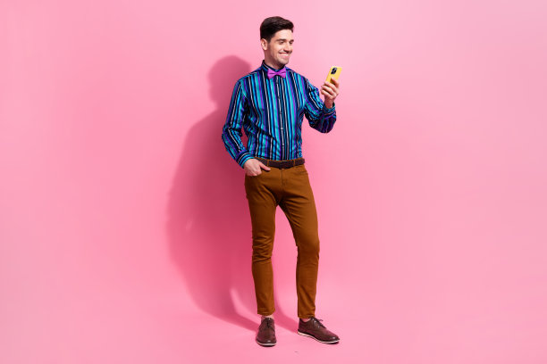 在粉红色的背景下，英俊的年轻有趣的积极的男人浏览智能手机的全身照片，拿着他的设备聊天图片下载