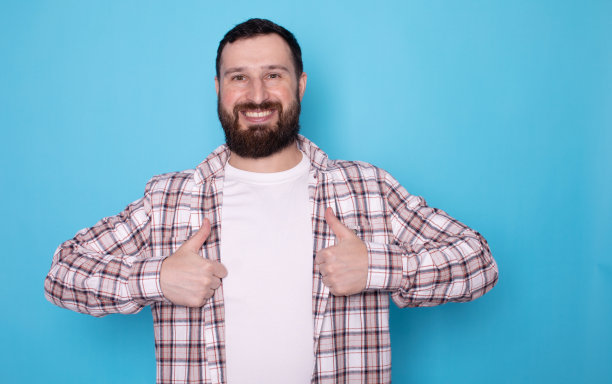 照片中，开朗英俊的大胡子男子露出獠牙，向你竖起大拇指，在蓝色背景上展示产品的质量。图片下载