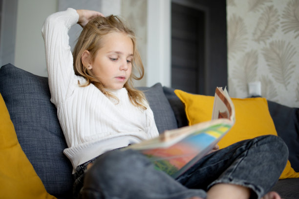 一个金发小女孩坐在家里的沙发上，沉浸在一本有趣的书的迷人情节中。图片下载