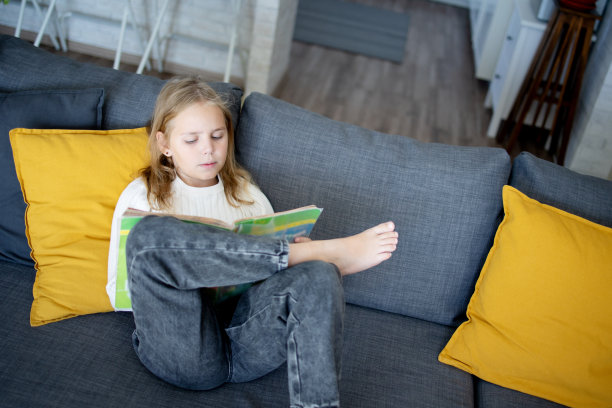 一个可爱的小女孩在家里舒适的环境中，按照低年级学生的课程，读着聪明而有趣的书。图片下载