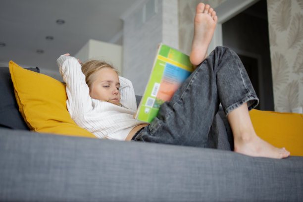 一个金发的低年级学前班女孩坐在家里的沙发上，热情地沉浸在一本令人兴奋的书的内容中。图片下载