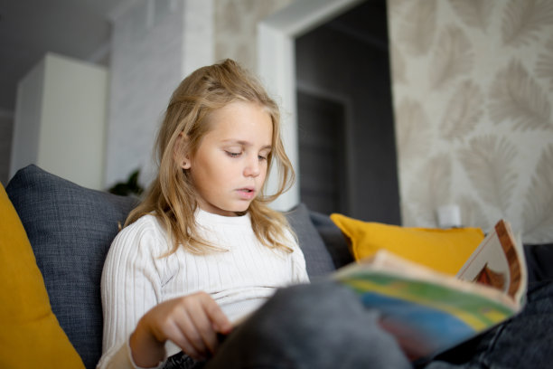 一个金发浓密的小女孩坐在舒适的沙发上，沉浸在书的迷人世界里。图片下载