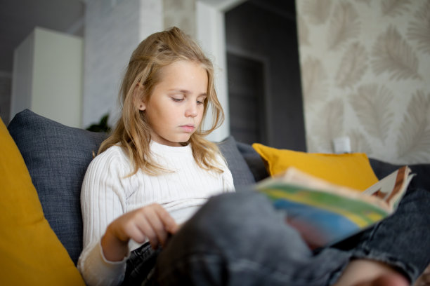 在家里的沙发上，一个穿着牛仔裤的金发小女孩正在读一本有趣的书。图片下载