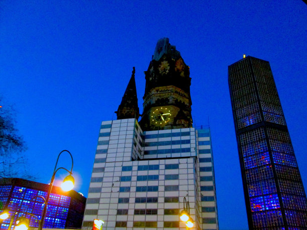 柏林蔚蓝的夜晚被摧毁的福音教堂钟楼和现代霓虹灯照亮的建筑物与路灯和光秃秃的树枝剪影图片下载