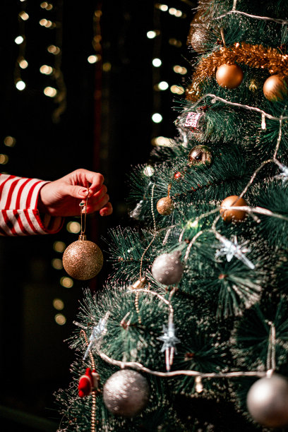 女人用闪亮的金色装饰物装饰圣诞树。为圣诞节做准备。现代闪闪发光的装饰在树的背景灯。Happy Holidays库存照片图片下载