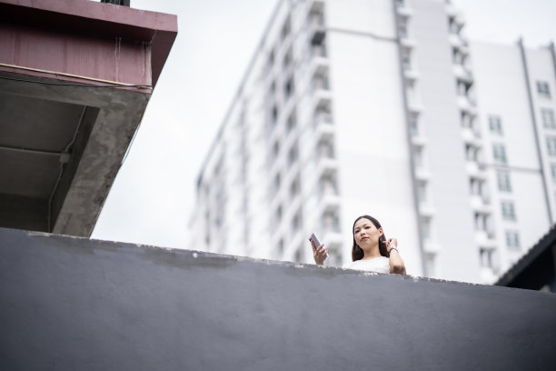 头部照片和最小的女人，建筑和天空的一部分，年轻的亚洲女人穿着白色t恤，拿着智能手机，自信地看着镜头，对城市的生活方式充满信心。图片下载
