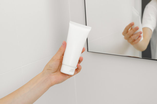 浴室里镜子的背景上，一个年轻女子的手拿着一管白色的奶油或牙膏模型。脸部和身体护理的概念，美容产品。图片为您的设计图片下载