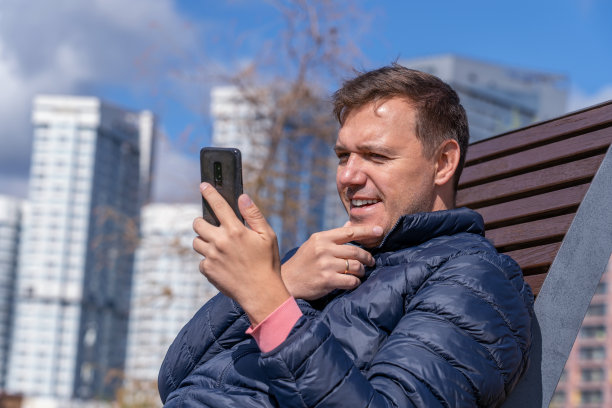 秋天，一名成熟的男子躺在城市公园的躺椅上，手放在下巴上，用智能手机开心地笑着。一个成熟的男人坐在公园的木凳上发短信图片下载