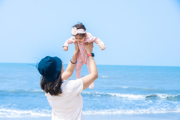 在热带海滩上，一位年轻的印度母亲将她可爱的小婴儿抛向天空。妈妈和小女儿在沙滩上的照片。图片下载