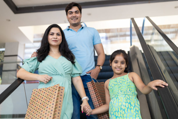 幸福的印度一家三口在商场里拎着纸袋，手握扶梯扶手和扶梯。购物的概念图片下载