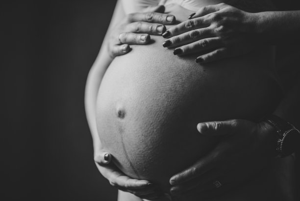 黑白照片孕妇，可爱的帅哥在家里抱着肚子。等待宝贝。九个月的特写。爱的夫妻。丈夫把手放在怀孕妻子的肚子上。亲子关系的概念。图片下载