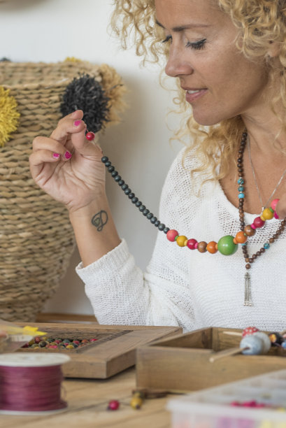 年轻的成年妇女在家里的桌子上制作珠子珠宝-现代家庭工作与珠子和更便宜的珠宝-网上商店的概念与创意的人图片下载