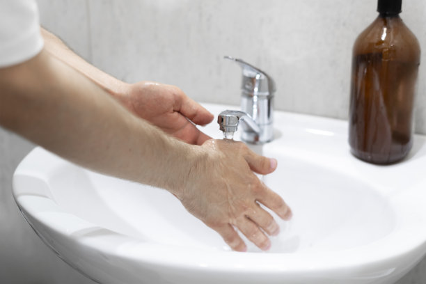 这张特写照片捕捉了洗手的动作，重点是双手在白色水槽旁边的水龙头下一起洗手，旁边是一个棕色的肥皂机。图片下载