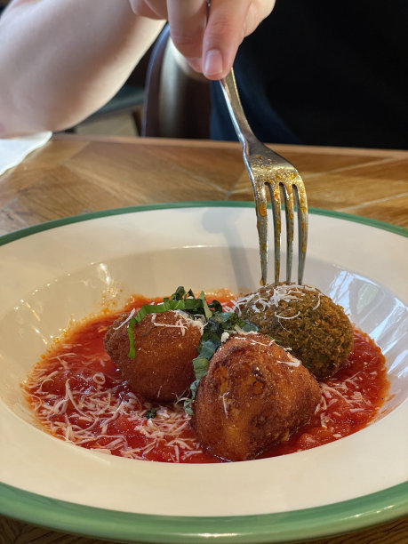 特写图片中，无法辨认的人用金属叉子吃阿兰西尼饭团，白色的碗里装着三个，阿兰西尼饭团配上番茄酱，上面撒上磨碎的帕尔马干酪，餐厅设置背景，重点放在前景图片下载