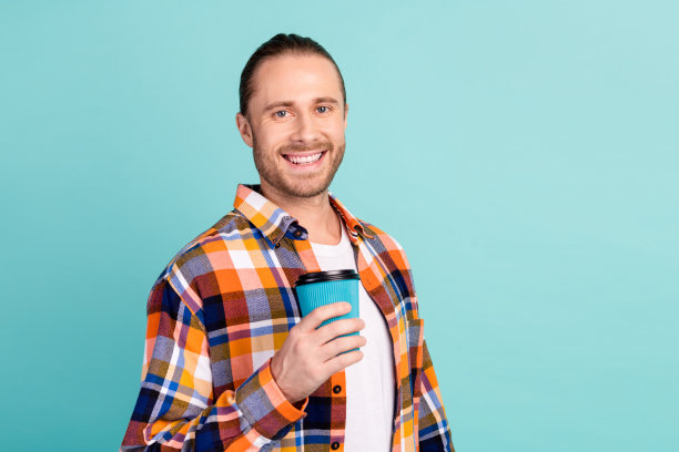 照片上的男人满脸胡须，穿着格子衬衫，拿着外带的咖啡杯，在蓝绿色的背景上显得很满足图片下载