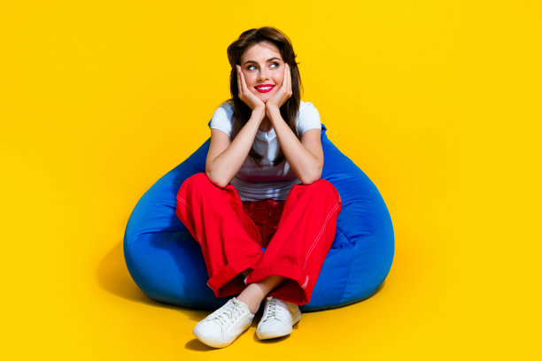 全身照片正面闪亮的女人穿着白色t恤坐在豆袋手臂脸颊看起来空空间孤立的黄色背景图片下载