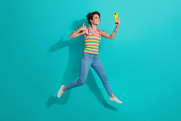 迷人的运动女孩的全尺寸照片跳拿智能手机自拍显示拇指向上孤立的蓝绿色背景图片下载